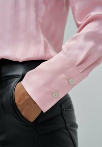 Rosa gestreiftes Bluse mit langen Ärmeln und geknöpften Manschetten, kombiniert mit schwarzen Lederhosen. Der Stoff wirkt glatt und leicht glänzend.