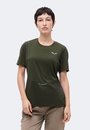 Femeie cu păr scurt și închis la culoare, purtând o cămașă verde măsliniu cu mâneci scurte și pantaloni bej, stând în fața unui fundal simplu și deschis la culoare.