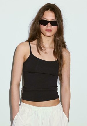 Młoda kobieta z długimi brązowymi włosami, nosząca czarne okulary przeciwsłoneczne, czarny top na cienkich ramiączkach oraz białe spodnie z elastycznym pasem, stoi na tle jednolitego tła.