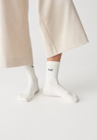 Weiße gerippte Socken mit umgeschlagenem Saum, auf der Seite mit dem Wort "hej!" in Schwarz bedruckt. Getragen mit hellbeigen weit geschnittenen Hosen.