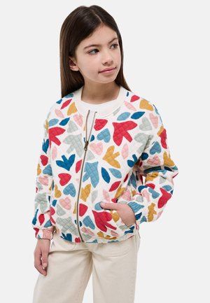 Fille aux longs cheveux bruns portant une veste matelassée blanche avec un motif abstrait de feuilles colorées et un pantalon crème, mains dans les poches, regardant vers la droite.