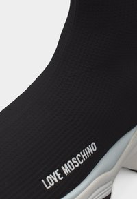 Μαύρο sneaker σε στυλ κάλτσας με υφή, παχιά λευκή σόλα και λευκό λογότυπο «LOVE MOSCHINO» στο πλάι κοντά στη σόλα.