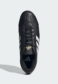 Černá sportovní obuv s bílými pruhy, texturovaný kožený svršek, zaoblená špička a černé tkaničky. Zlato logo na jazyku.