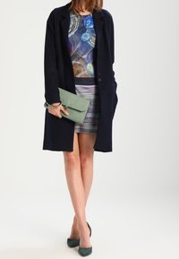 Cappotto lungo blu navy sopra una camicia multicolore e una gonna a righe, abbinati a una pochette verde chiaro e tacchi verde scuro. Texture lisce evidenti.