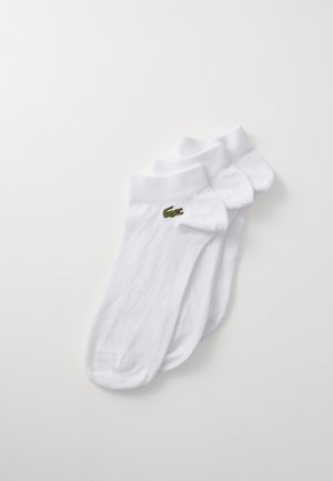 Chaussettes blanches en coton, avec une bordure côtelée et un logo crocodile brodé vert. Texture lisse, trois paires disposées soigneusement.