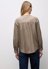 Camicia a maniche lunghe color beige con scollatura arricciata, dettaglio arricciato sul retro e polsini con bottoni, abbinata a jeans blu scuro.