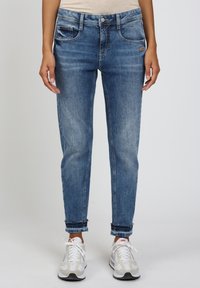 Person in blauen, ausgewaschenen Jeans mit hochgekrempelten Säumen und weißen Nike-Sneakern, der vor einem schlichten, hellen Hintergrund steht.