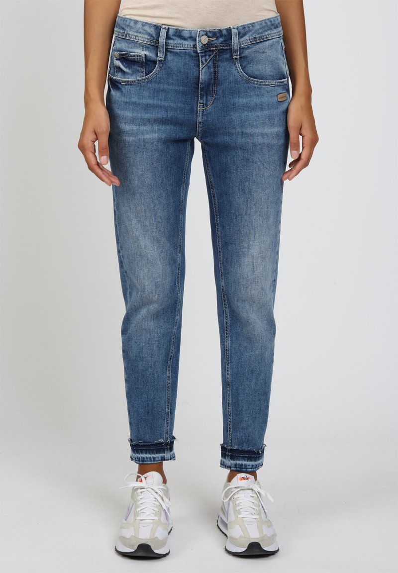 Person in blauen, ausgewaschenen Jeans mit hochgekrempelten Säumen und weißen Nike-Sneakern, der vor einem schlichten, hellen Hintergrund steht.