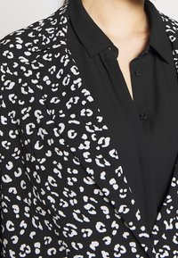 Blazer noir à imprimé léopard blanc, avec un tissu texturé et un col classique. En dessous, une chemise noire à boutons.