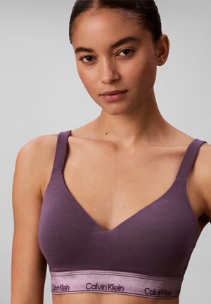 Femme portant un bralette violet Calvin Klein avec larges bretelles et bande élastique à logo, sur fond gris uni.
