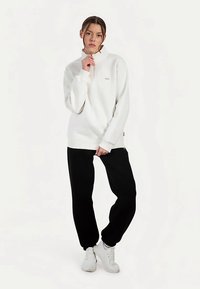Biała bluza z wysokim kołnierzem i długimi rękawami, zestawiona z czarnymi joggerami i białymi trampkami. Materiał wydaje się miękki i gładki.