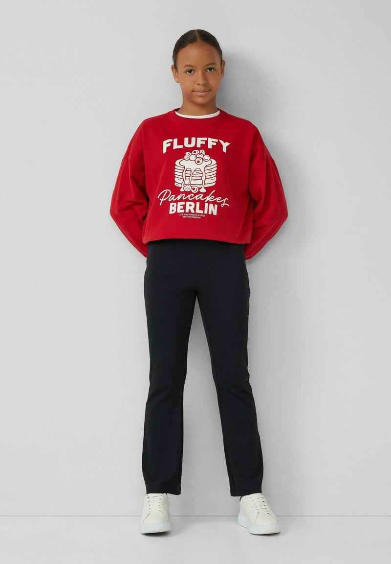 Felpa rossa con grafica bianca che raffigura pancake e il testo "Fluffy Pancakes Berlin", abbinata a pantaloni neri aderenti e sneakers bianche.