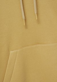 Gros plan sur un tissu de sweat à capuche marron clair montrant des cordons avec embouts métalliques dorés et une poche kangourou à l'avant.