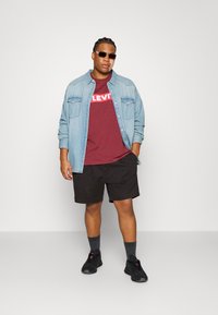 Denimskjorte over en mahogni Levi's t-shirt, sorte shorts, sorte sneakers og grå sokker. Skjorten har knapdetaljer og lommer.