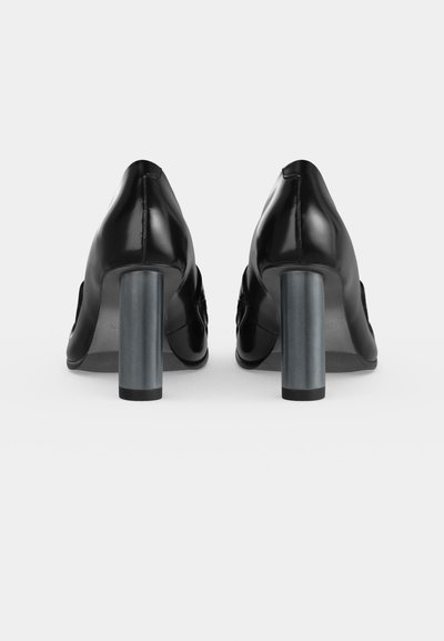 Paire de chaussures à talons hauts en cuir noir brillant montrées de dos avec des talons cylindriques épais sur un fond clair uni.