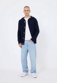 Férfi sötétkék gombos kardigánban, kapucnival, fehér pólóban, világoskék farmernadrágban és fehér sportcipőben áll, az egyik kezét a zsebében tartva.