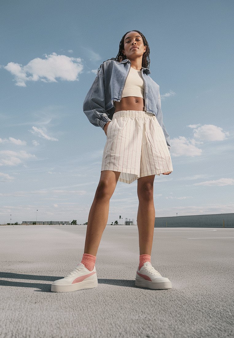 Jeans-Crop-Jacke, weiße Shorts mit feinen Streifen, weiße und rosa Sneakers mit strukturierten Sohlen und rosa Socken; vor einem klaren blauen Himmel.