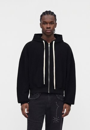 Jonge man met gevlochten haar draagt een zwarte zip-up hoodie met crèmekleurige trekkoorden en zwarte jeans met studs op één been.