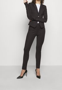 Schwarzer taillierter Blazer mit Knopfakzenten, kombiniert mit eng anliegenden schwarzen Hosen und spitzen schwarzen High Heels. Glattes Gewebe mit schlankem Design.