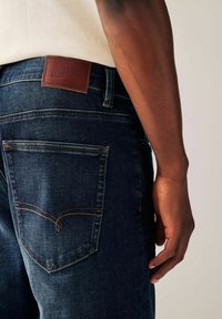 Dark denim jeans with orange stitching, back pocket detailing, and a brown leather label at the waistband showcasing "DENIM GOODS."