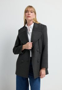 Abrigo de lana gris de doble botonadura con solapas anchas, dos grandes botones y bolsillos laterales, combinado con una camisa blanca y jeans azules.