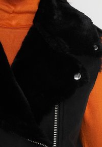 Svart fuskpälskrage på en svart läderjacka, med silverknappar och en metallblixtlås, lager på lager över en orange polotröja.