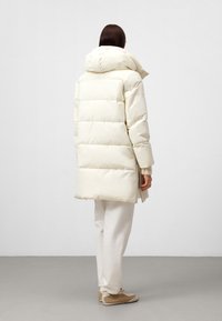 Kvinna iklädd en lång, off-white, quiltad pufferjacka med huva, vita byxor och beige sneakers, som står mot en enkel ljus bakgrund.