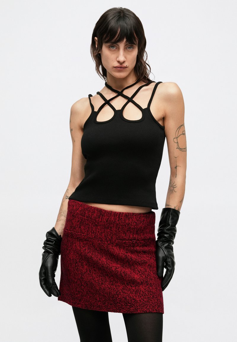 Black ribbed crop top with crisscross straps and round cutouts, paired with a red textured mini skirt. Model wears black leather gloves.