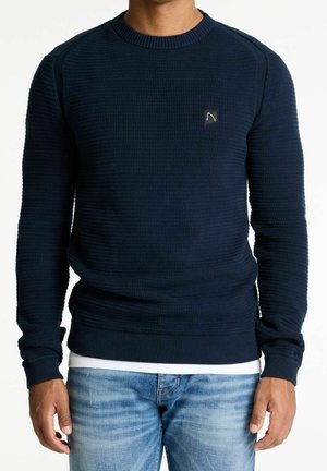 Uomo che indossa un maglione lavorato a maglia di colore blu scuro sopra una camicia bianca e jeans azzurri, in piedi con le braccia lungo i fianchi.