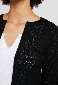 Svart, stickad cardigan med ett strukturerat diamantmönster och öppen väv. Den har en rund halsringning och är lager på lager över en vit topp.