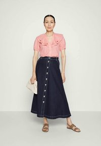 Mulher vestindo uma blusa rosa de mangas curtas com babados, uma saia ajustada em A de botão escuro, sandálias bege, segurando uma clutch bege, em pé sobre um fundo claro.