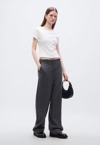 Λευκό κοντομάνικο τοπ με κυματιστές άκρες, συνδυασμένο με φαρδιά γκρι παντελόνια με ρίγες. Το μοντέλο κρατάει μια μικρή μαύρη τσάντα.