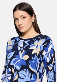 Femme aux longs cheveux bruns portant un pull bleu marine avec de grands motifs floraux dans des nuances de bleu, beige et blanc, sur un fond blanc.