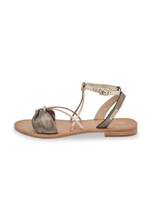 Sandalia plana con punta metálica dorada, correas de cuero, cierre de hebilla y una suela de color marrón claro. Presenta un diseño anudado en la punta.