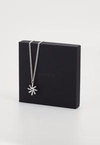 Colar de corrente prateada com um pendente em forma de flor, apresentando um centro escuro, exibido numa caixa retangular preta com um logotipo subtil.