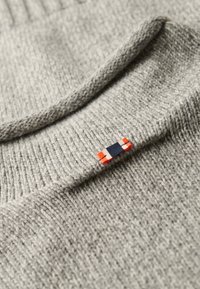 Grau gestrickter Pullover mit rundem Ausschnitt, der ein kleines, lebendiges Patch aus roten, marineblauen und weißen Stoff an der Kragennaht aufweist.