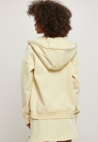Sudadera oversized de color amarillo pálido con una gran capucha, puños acanalados y un dobladillo trasero más largo, hecha de suave tela de algodón.