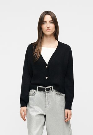 Femme portant un cardigan noir à col en V avec des boutons en forme de cœur sur un haut blanc, assorti d'un jean taille haute gris clair et d'une ceinture noire.