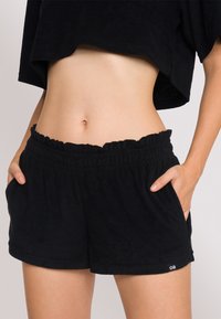 Pantaloni corti in spugna nera con vita elastica arricciata, tasche laterali e una piccola etichetta con logo. Tessuto morbido e vestibilità rilassata.