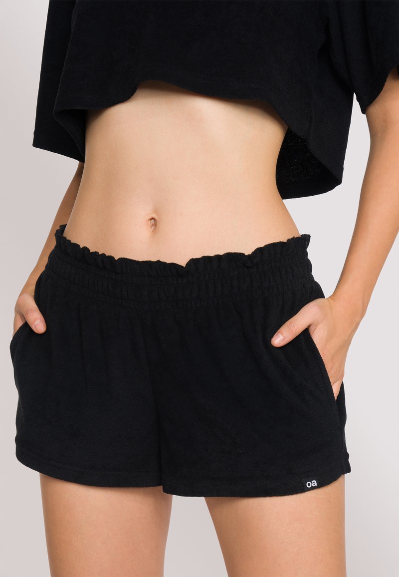 Pantaloni corti in spugna nera con vita elastica arricciata, tasche laterali e una piccola etichetta con logo. Tessuto morbido e vestibilità rilassata.