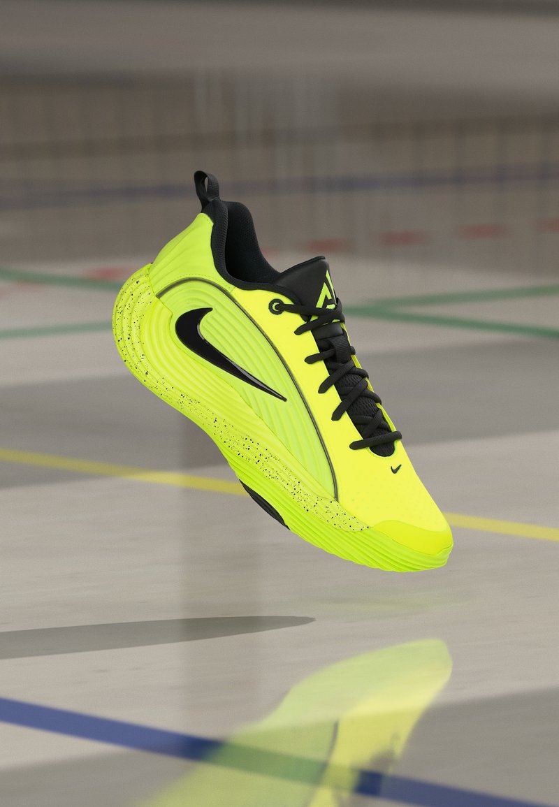Baskets athlétiques jaune vif et noir avec logo swoosh noir, en l’air au-dessus d’un terrain de sport intérieur poli avec des lignes de délimitation colorées.