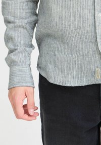 Striped blue and white button-up shirt made of lightweight fabric, with a cuffed sleeve and an additional tag detail near the hem. Black pants are visible.