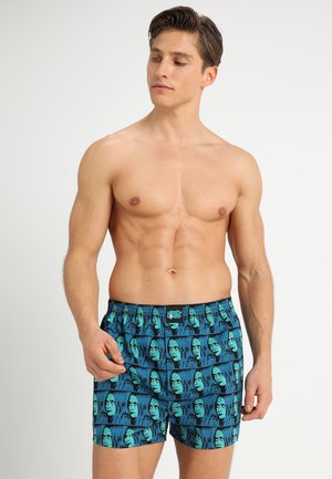 Blaue Boxershorts mit einem wiederholten Grafikmuster aus grünen und schwarzen Gesichtern, aus leichtem Stoff gefertigt und mit einem elastischen Bund.