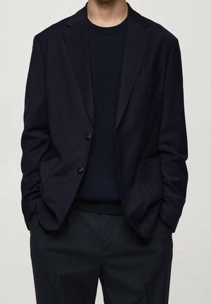 Mand iført en mørk marineblazer over en sort crewneck-shirt med matchende mørke bukser, hænderne i lommerne, neutral baggrund.