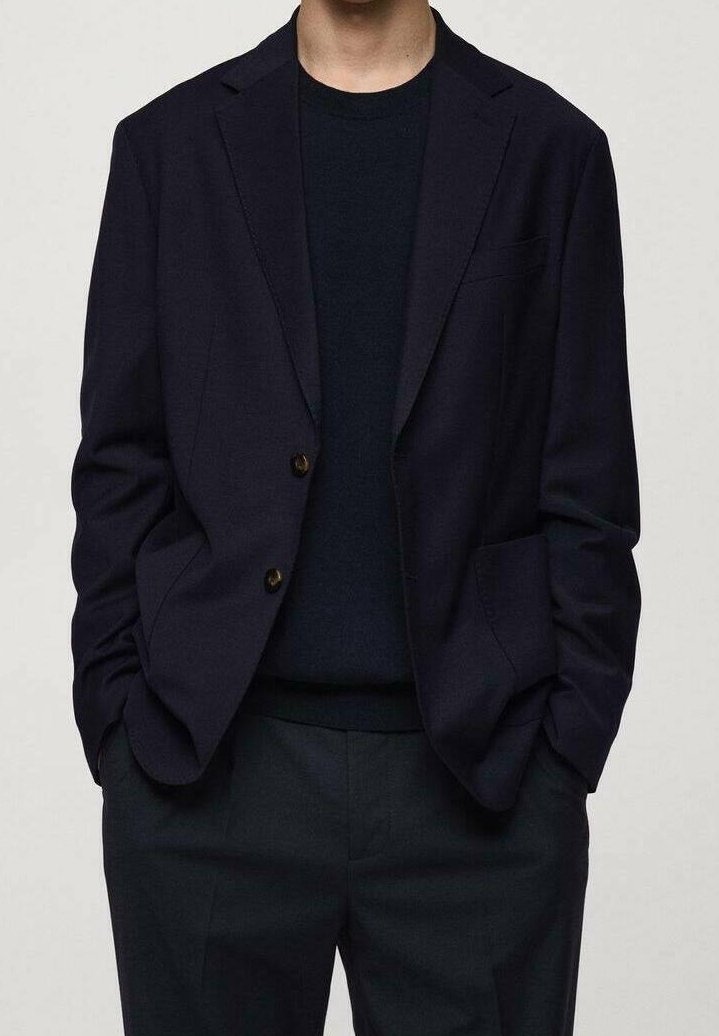 Homme portant un blazer bleu marine foncé sur un t-shirt noir à col rond avec un pantalon foncé assorti, mains dans les poches, fond neutre.