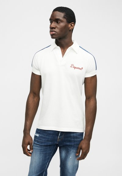 Homme portant un polo blanc à manches courtes avec le logo "Dsquared²" et un jean bleu, debout devant un fond clair uni, regardant vers la gauche.