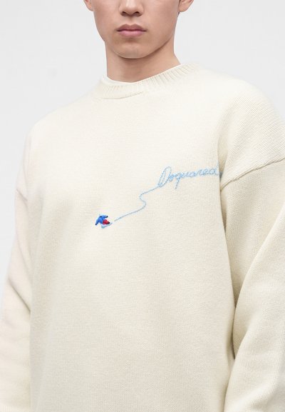 Homme portant un pull écru avec un texte cursif bleu clair et un petit motif brodé bleu et rouge sur la poitrine.