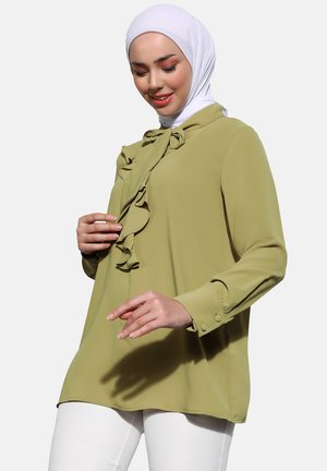 Olive green blouse with a ruffled front detail and buttoned cuffs, made of lightweight fabric. Worn over white pants.