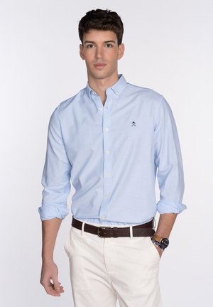 Camisa de botones azul claro con mangas enrolladas, que presenta un pequeño logo bordado en el pecho, combinada con pantalones blancos y un cinturón marrón oscuro.