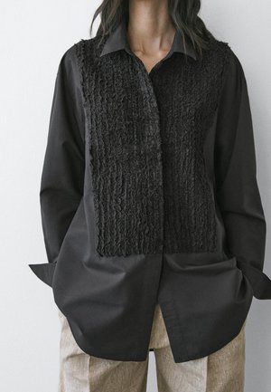 Personne portant une chemise noire à manches longues avec un panneau avant texturé et un pantalon beige, debout les mains dans les poches devant un fond uni.